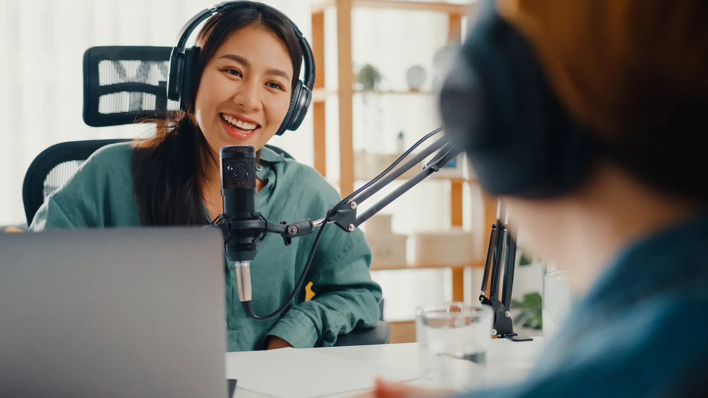 A host in headphones smiling at the person she is interviewing across a table, a condenser microphone between them and a laptop open in front of her.