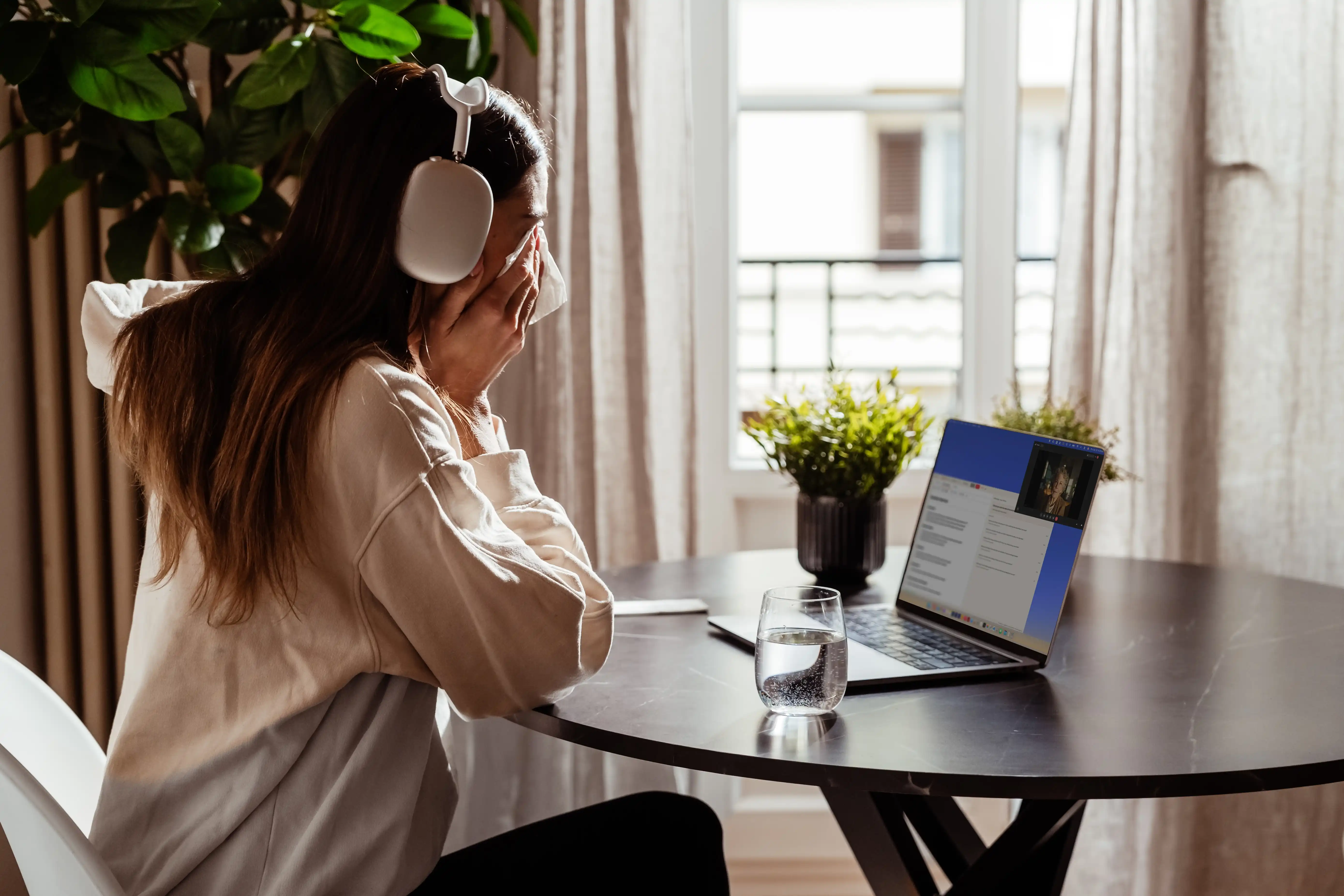 A young woman on a video call at her kitchen table, headphones on, overcome by what her grandmother is telling her on the screen.